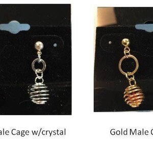 Puede incluir: Dos pares de pendientes con un dise&ntilde;o de jaula. Los pendientes de la izquierda son de plata con un cristal transparente en la jaula. Los pendientes de la derecha son de oro con un cristal rojo en la jaula. Los pendientes est&aacute;n etiquetados como "Silver Male Cage w/crystal" y "Gold Male Cage w/crystal".