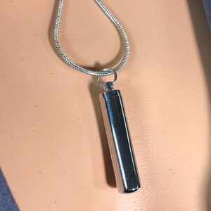May include: A silver chain necklace with a cylindrical pendant. The pendant is smooth and shiny, and it has a rounded top and bottom.