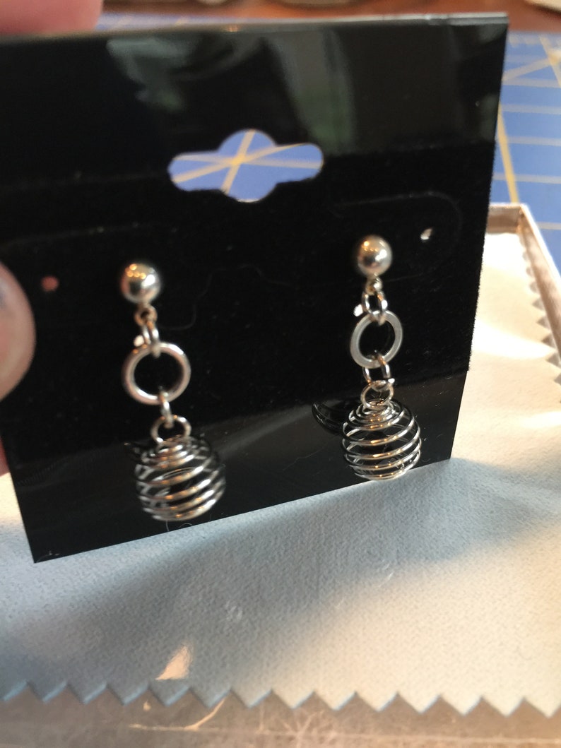 Puede incluir: Un par de pendientes de plata con un dise&ntilde;o de jaula colgante. Cada pendiente tiene una peque&ntilde;a bola de plata en la parte superior, seguida de un peque&ntilde;o aro de plata y luego una jaula colgante hecha de alambre de plata.