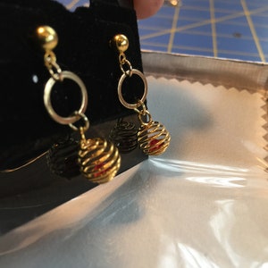 Puede incluir: Pendientes de oro con una peque&ntilde;a perla roja dentro de una jaula dorada. Los pendientes tienen un peque&ntilde;o aro de oro por encima de la jaula.