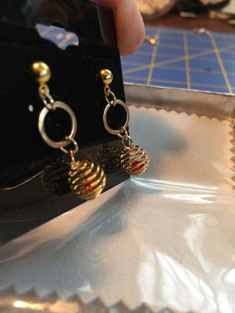 Puede incluir: Pendientes de oro con una peque&ntilde;a perla roja dentro de una jaula dorada. Los pendientes tienen un peque&ntilde;o aro de oro por encima de la jaula.