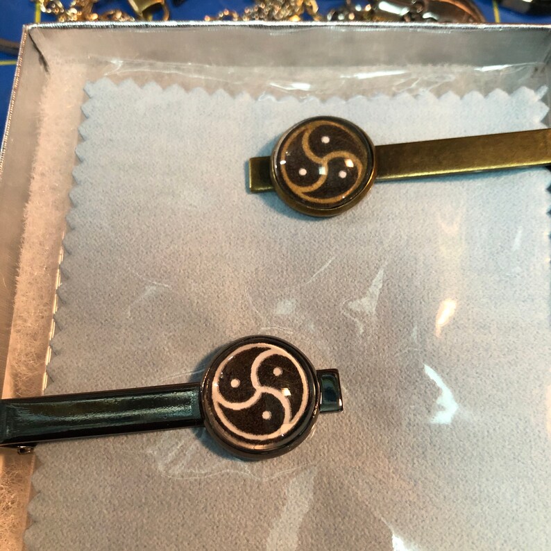 May include: Two tie clips with circular designs. One clip is gold-toned with a brown and white symbol. The other is black with a similar design. Both are displayed in a box.