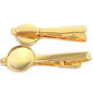May include: Two gold-colored tie clips. Each clip has a rectangular bar with a hinged clasp and a circular bezel. The clips are made of shiny, polished metal. The clasps have a ridged design for grip.