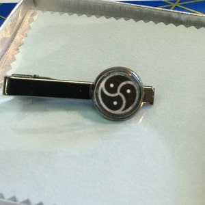 May include: A black metal tie clip with a circular emblem featuring a three-lobed design. The tie clip is rectangular and rests on a light blue surface inside a silver box. The emblem is black and white.
