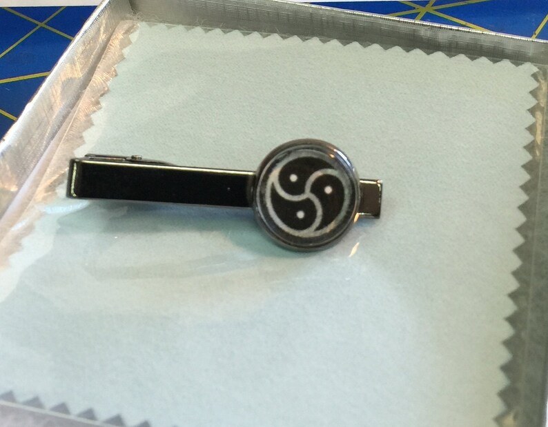 May include: A black metal tie clip with a circular emblem featuring a three-lobed design. The tie clip is rectangular and rests on a light blue surface inside a silver box. The emblem is black and white.