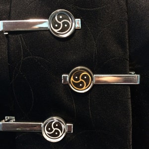 May include: Three silver-toned tie clips with circular designs. Two clips feature a black and white symbol, while the center clip has a black and gold design. The clips are displayed on a black fabric background.