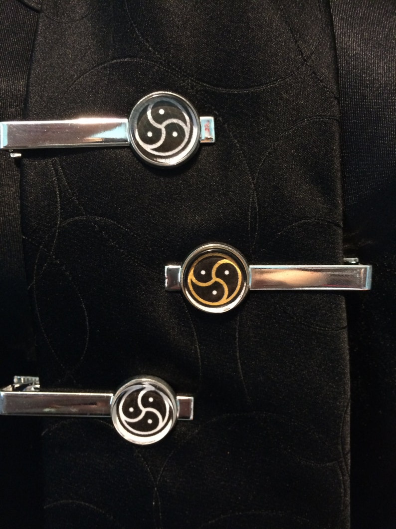 May include: Three silver-toned tie clips with circular designs. Two clips feature a black and white symbol, while the center clip has a black and gold design. The clips are displayed on a black fabric background.