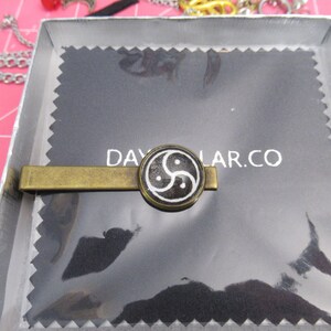 May include: A gold-toned tie clip with a circular black and white design. The design features a stylized symbol. The tie clip is presented in a silver box on a black background with the text DAYLAR.CO.