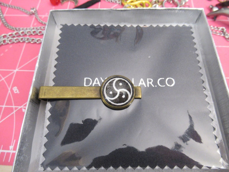 May include: A gold-toned tie clip with a circular black and white design. The design features a stylized symbol. The tie clip is presented in a silver box on a black background with the text DAYLAR.CO.