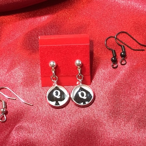May include: Silver-toned earrings featuring a black spade design with a white 'Q' in the center. The earrings are displayed on a red surface, with additional earring hooks visible. The earrings are a playful accessory.
