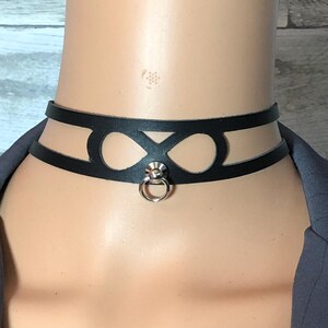 May include: Black leather choker necklace with a cut-out design featuring two oval shapes and a central X. A silver-colored ring hangs from the center. The choker is worn around the neck.