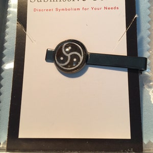 May include: A black metal tie clip with a circular emblem featuring a white and black symbol. The text "Submissive Gear" and "Discreet Symbolism for Your Needs" is visible on the packaging.