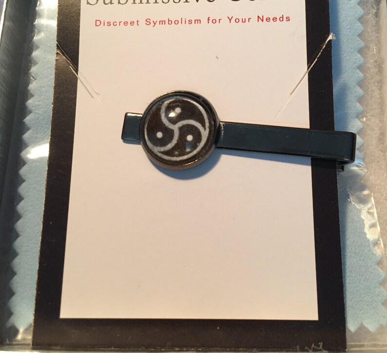 May include: A black metal tie clip with a circular emblem featuring a white and black symbol. The text "Submissive Gear" and "Discreet Symbolism for Your Needs" is visible on the packaging.