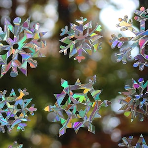May include: A collection of 12 iridescent snowflake window clings. The snowflakes are made of a clear, reflective material that catches the light and creates a shimmering effect. They are perfect for adding a touch of sparkle to your home during the holiday season.