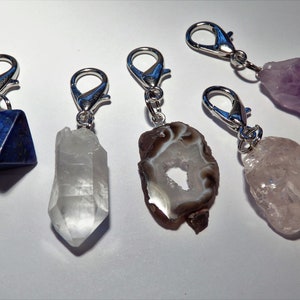 May include: Five crystal charms with silver clasps. The charms are a clear quartz point, a brown agate with a hole in the center, a purple amethyst, a pink rose quartz, and a blue lapis lazuli pyramid.