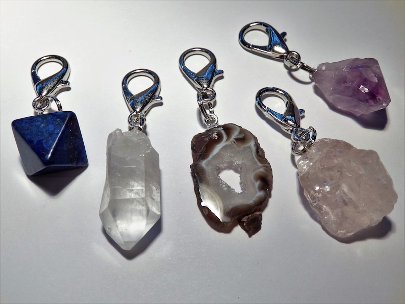 May include: Five crystal charms with silver clasps. The charms are a clear quartz point, a brown agate with a hole in the center, a purple amethyst, a pink rose quartz, and a blue lapis lazuli pyramid.