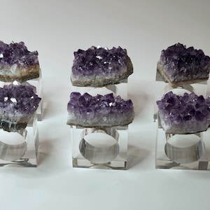 May include: Six amethyst crystal napkin rings on clear acrylic bases. The amethyst crystals are a deep purple color and have a natural, rough texture.