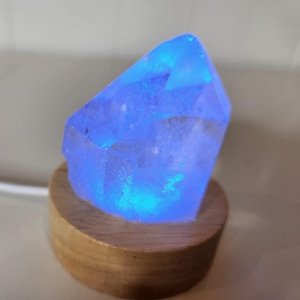May include: A clear crystal shaped like a point, illuminated from within with a bright blue light. The crystal is sitting on a wooden base.