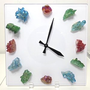 May include: Square white clock with black hands and colorful crystal accents. The clock face features various clusters of blue, green, red, and iridescent crystals, arranged to mark the hours. The clock is supported by clear plastic stands.