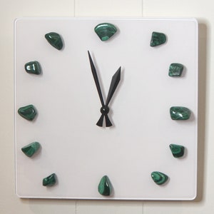 May include: Square white wall clock with black hands and green malachite stone hour markers. The clock has a minimalist design, with the stones arranged around the clock face to indicate the hours.