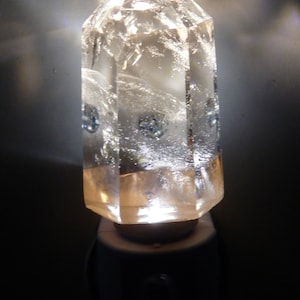 May include: A clear crystal shaped like a pyramid is illuminated from within, creating a warm glow. The crystal is mounted on a white base and plugged into an electrical outlet.