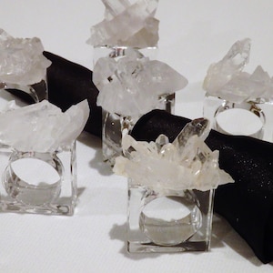 May include: Five clear crystal napkin rings with a cluster of quartz crystals on top. The rings are square and made of glass. The rings are sitting on a black napkin.