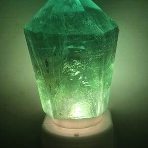 May include: A faceted, emerald-green crystal night light. The crystal is mounted on a white base with a soft, glowing light emanating from within. The crystal's surface has visible internal structures.