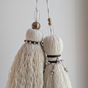May include: Two off-white decorative tassels with a textured, fringed design. Each tassel features a spherical top with a gold or clear bead accent and a string for hanging. The tassels are held up against a plain white background.