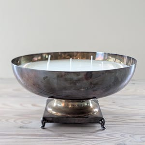 May include: A large, antique silver-toned bowl candle with multiple wicks. The candle sits on a square, silver-toned base with small feet. The candle's wax is white, and the bowl has a tarnished appearance.