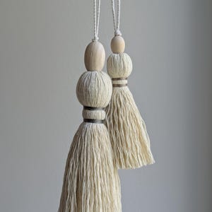 May include: Two off-white cotton tassels hanging from white cords. Each tassel features a smooth, light-colored wooden bead at the top, a round yarn-wrapped sphere, and a dark grey thread binding above the long, loose, fringed yarn skirt.
