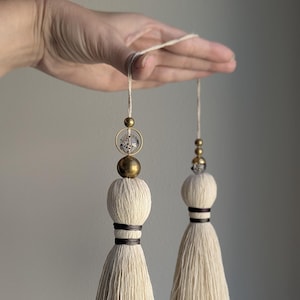 May include: Two off-white cotton tassels suspended from a single string. Each tassel features a rounded top section wrapped with dark brown thread, a large brass-toned bead, a small brass-toned bead, and a clear bead with black speckles.