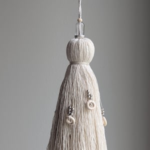 May include: A cream-colored fiber tassel hanging from a white cord. The top features a clear glass bead and a small wooden bead. The tassel is adorned with several silver-toned metallic beads and small, circular off-white rings attached to the strands.