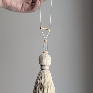 May include: Cream-colored tassel with a decorative wooden bead and a clear glass bead. The tassel is suspended from a white cord with a small gold-colored bar. The design is simple and elegant, suitable for home decor.
