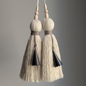 May include: Two off-white decorative tassels featuring thick yarn fringe, wrapped tops, and wooden beads on the hanging cords. Each tassel includes a small, dark brown, cone-shaped metal accent suspended from the center of the yarn strands.
