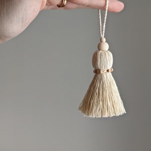 May include: A white tassel with a wooden bead at the top and a row of wooden beads around the middle. The tassel is hanging from a white string.