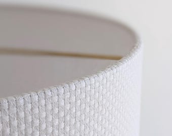 May include: A cylindrical lamp shade featuring a crisp white fabric exterior with a distinct woven basket-weave texture. The shade has a clean, structured edge and a smooth interior lining, showcasing the detailed grid pattern of the textile material.