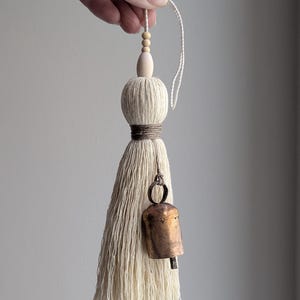 May include: Cream-colored tassel with a wooden bead accent and a small, aged bronze bell. The tassel is made of thick, textured yarn and is tied with a brown cord. A white string is attached for hanging.
