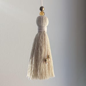 May include: A cream-colored tassel with a wrapped top and dangling fringe. The tassel is adorned with gold-colored beads and small gold-colored charms. It hangs from a thin, white string against a neutral background.