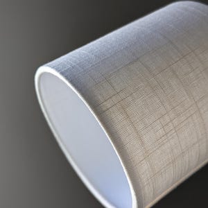 May include: A cylindrical lamp shade featuring a light off-white fabric with a subtle, intersecting grid pattern. The material has a visible woven texture, and the shade is open at the top and bottom, showing a smooth, solid white interior lining.
