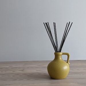 May include: A yellow ceramic diffuser vase with a handle, filled with black reeds. The vase sits on a wooden surface against a neutral background. The design is simple and modern, suitable for home fragrance.