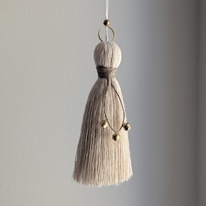 May include: A handmade, off-white tassel with a wrapped midsection and hanging gold-colored bells. The tassel is suspended by a white cord and features a small gold bead and ring at the top. The design is simple and elegant.