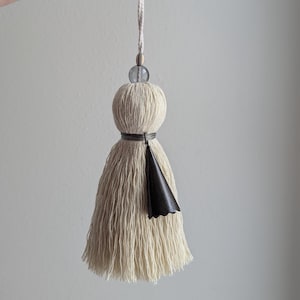 May include: Cream-colored tassel with a black leather accent and a clear bead. The tassel is suspended by a thin, off-white cord. The design features a rounded top and a flared, fringed bottom.
