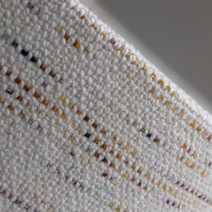 close up photo of woven fibers in mostly ivory with yellows, golds, reds, blues, and purples running through it