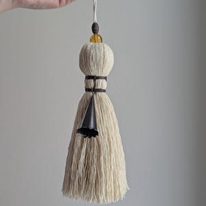 May include: Cream-colored tassel with a decorative design. The tassel features a bell-shaped metal accent and a wooden bead. The top has a small yellow bead and a wooden bead. The tassel is suspended by a natural-colored cord.