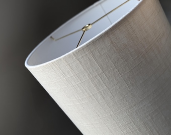 Puede incluir: Una pantalla de lámpara cilíndrica con un exterior de tela texturizada de color beige claro con un patrón de tejido cruzado visible. El interior está forrado con material blanco liso e incluye un soporte de metal dorado en la parte superior.