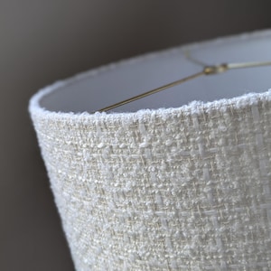 May include: Close-up of a cream-colored lampshade with a textured, woven fabric. The lampshade has a white interior and is supported by a gold-colored metal frame. The fabric has a subtle, light-reflecting quality.