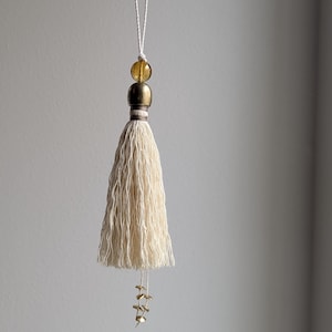 May include: Cream-colored tassel with a brass-colored bead and a golden-yellow bead at the top, suspended by a white string. Small brass-colored charms dangle from the bottom of the tassel. The tassel is made of a soft, textured material.
