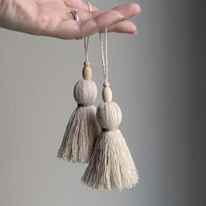 May include: Two beige cotton tassels featuring a rounded top section and a flared fringe base. Each tassel is accented with a smooth, light-colored wooden bead at the top and is suspended by a matching twisted cotton cord loop for hanging.