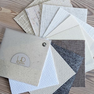 Ud Form fabric sample book on light wood table showing custom lamp shade fabrics in whites, creams, naturals, brown, grey, and multicolor.