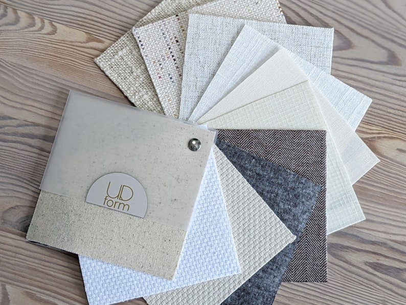 Ud Form fabric sample book on light wood table showing custom lamp shade fabrics in whites, creams, naturals, brown, grey, and multicolor.